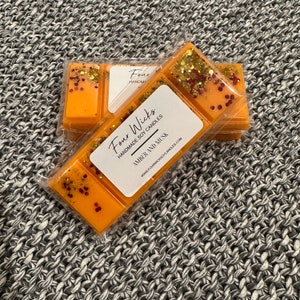 May include: Three wax melts in clear packaging with a label that reads "Four Wicks Handmade Soy Candles Amber and Musk www.fourwickssoycandles.com". The wax melts are orange with red and gold glitter.