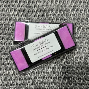Puede incluir: Dos ceras aromáticas rectangulares en envase transparente. Cada cera tiene un color púrpura oscuro y está dividida en tres secciones. La etiqueta dice "Four Wicks Handmade Soy Candles" y "Midnight Romance Type."