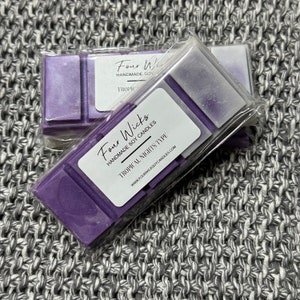 Puede incluir: Dos fundidos de cera de soja púrpura empaquetados en plástico transparente. Los fundidos de cera están etiquetados como "Four Wicks Handmade Soy Candles" y "Tropical Nights Type".
