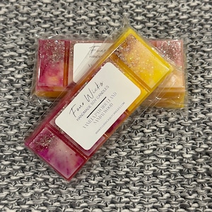 Puede incluir: Dos fundentes de cera en un envase transparente. Uno es rosa y amarillo con purpurina, el otro es amarillo con purpurina. La etiqueta dice "Four Wicks Handmade Soy Candles Vanilla Patchouli and Sandalwood www.fourwicksoycandles.com"