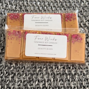 May include: Two wax melt bars in clear packaging. Each bar is divided into four sections, with a light orange colour and pink glitter. The labels read "Four Wicks Handmade Soy Candles" and "Dragons Blood".