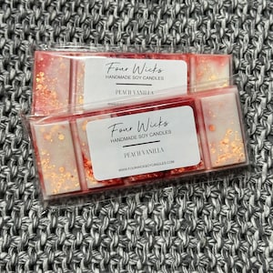 Puede incluir: Dos fundidos de cera en un envase transparente con un diseño rojo y blanco. Los fundidos de cera están etiquetados como "Four Wicks Handmade Soy Candles Peach Vanilla" e incluyen la dirección del sitio web "www.fourwicksoycandles.com".