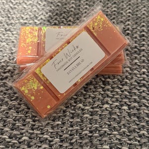 May include: A stack of four-wick soy wax melts in clear plastic packaging. The top melt is labelled "Four Wicks Handmade Soy Candles Banana Bread www.fourwicksoycandles.com". The melts are a light peach colour with gold glitter.