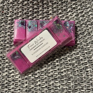 May include: A pink and purple wax melt with glitter, packaged in clear plastic. The label reads "Four Wicks Handmade Soy Candles Watermelon Lemonade www.fourwicksoycandles.com".