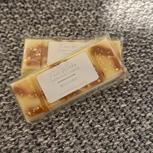 May include: A white wax melt bar with gold flecks and a brown swirl design. The bar is labeled "Four Wick's Handmade Soy Candles" and "Banana Split".