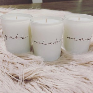Puede incluir: Tres velas votivas blancas con los nombres "gabe", "mich" y "max" escritos a mano. Las velas están en recipientes de vidrio esmerilado y se colocan sobre una superficie suave y clara.