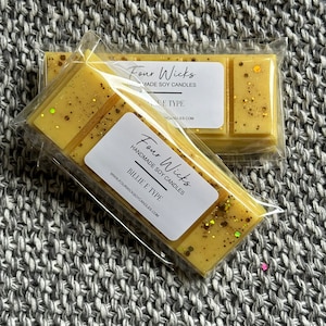 May include: Two wax melts in clear packaging. The wax melts are a light yellow color with gold glitter. The packaging has a white label with the text "Four Wicks Handmade Soy Candles Billie E Type www.fourwicksoycandles.com"
