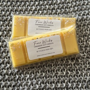May include: Two buttered popcorn scented soy wax melts in clear packaging. Each melt is a light yellow colour with gold and white glitter accents. The label reads "Four Wicks Handmade Soy Candles, Buttered Popcorn."