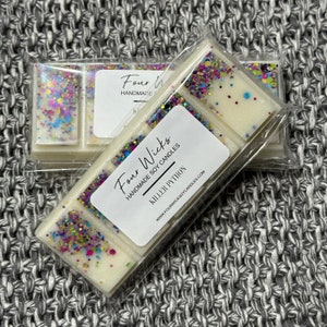 Puede incluir: Un derretimiento de cera blanco con purpurina colorida, empaquetado en plástico transparente. La etiqueta dice "Four Wicks Handmade Soy Candles" y "Killer Python" con la dirección del sitio web "www.fourwickssoyscandles.com".