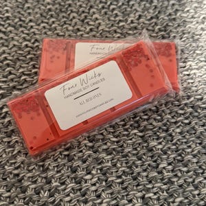 May include: Two red wax melt bars in clear packaging. The bars are labelled "Four Wicks Handmade Soy Candles" and "All Red Ones." The wax melts have red glitter accents. The background is a grey knitted fabric.