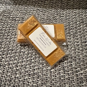 May include: A close-up of a wax melt bar with a white label that reads "Four Wicks Handmade Soy Candles Hot Baked Apple Pie" and a website address. The wax melt bar is a light brown colour with gold flecks.