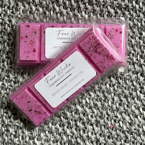 Può includere: Un fondente di cera di soia rosa con glitter in una confezione di plastica trasparente. L'etichetta riporta "Four Wicks Handmade Soy Candles Britney Spears Fantasy Type" e include un indirizzo di sito web.