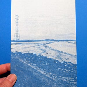 May include: A cyanotype print on paper featuring a landscape scene with a tall electrical transmission tower on the left, power lines stretching across the sky, and a winding river flowing through a grassy field under a pale horizon.
