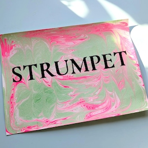 May include: A rectangular card with a marbled design in shades of pink and green. The word "STRUMPET" is printed in bold black letters across the center of the card. The card is on a light blue surface.