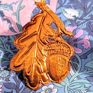 May include: A metallic copper-coloured ornament shaped like an oak leaf and an acorn attached to a small stem. The surface features embossed textures and details on the leaf veins and the acorn cap. A thin white string is attached to the top loop.