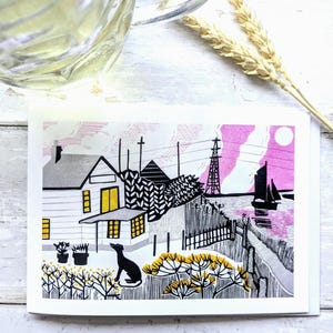 May include: A greeting card featuring a linocut print of a coastal scene. The artwork depicts a house, a dog, and a sailboat under a pink sky. The card is on a white background with a glass and wheat stalks.