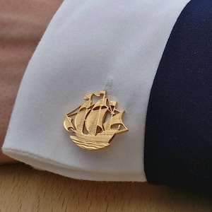 Può includere: Gemelli in oro con un designo di nave dettagliato. I gemelli sono su un polsino di camicia bianca con una giacca di abito blu scuro.