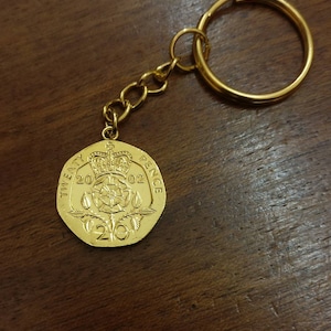 May include: Gold-coloured keychain with a 20 pence coin from the United Kingdom. The coin features a royal crest and the year 2002.