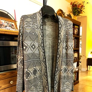 May include: A black, white, and tan open-front cardigan with a geometric pattern and fringe trim. The cardigan is displayed on a clothing rack. The design features diamond and linear shapes.
