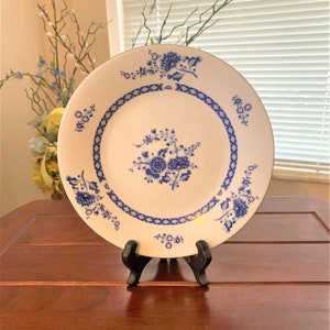May include: A white ceramic plate with a blue and white floral design. The plate has a decorative border with a diamond pattern. The plate is on a black stand.