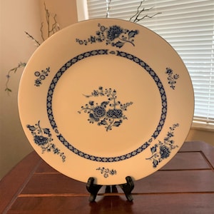 May include: A white ceramic plate with a blue and white floral design. The plate has a decorative border with a diamond pattern. The plate is on a wooden stand.