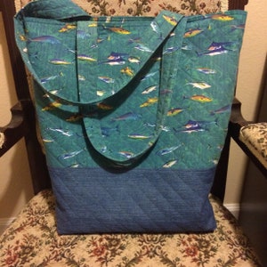 May include: A blue tote bag with a quilted design. The bag features a pattern of colorful fish on a teal background. The bottom of the bag is a solid blue color.