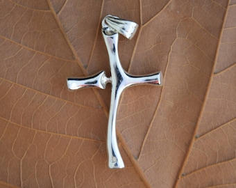 May include: A silver cross pendant with a distinctive, curved form. The cross is set against a large, dried brown leaf, with a textured wooden backdrop. The pendant has a polished finish.