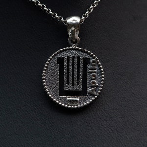 Puede incluir: Collar con colgante de plata con un dije redondo de plata texturizado que presenta una lira y la palabra "Apollo" inscrita alrededor del borde. El dije también tiene el número "925" estampado en él.