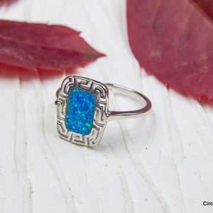 Op de afbeelding: Een zilveren ring met een blauwe opaal-steen gezet in een geometrisch ontwerp. De ring ligt op een wit houten oppervlak met een rood blad op de achtergrond.