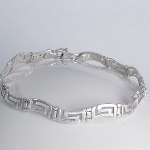 Könnte beinhalten: Ein silbernes Armband mit geometrischem Muster. Das Armband besteht aus ineinandergreifenden Quadraten und Rechtecken.