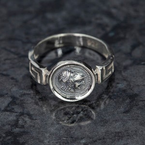 Può includere: Anello d'argento con un designo rotondo e dettagliato che presenta il profilo di una persona con un elmo. La fascia dell'anello ha un motivo a chiave greca.