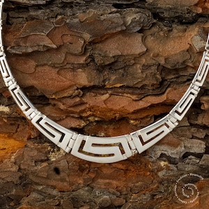 May include: A silver necklace with a geometric pattern, resembling a Greek key design. The necklace is photographed on a textured wooden surface.