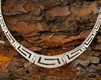 May include: A silver necklace with a geometric pattern, resembling a Greek key design. The necklace is photographed on a textured wooden surface.
