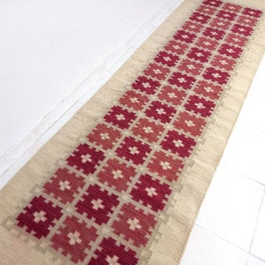 May include: A long, rectangular rug with a cream background and a geometric pattern of red and pink squares. The rug is woven with a traditional design.