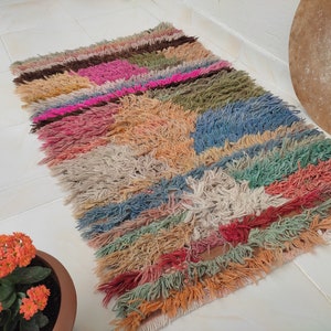 May include: A colorful, handwoven rug with a geometric pattern in shades of pink, blue, green, orange, and brown. The rug has a fringed edge and is made of soft, fluffy fibers.