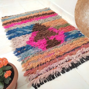 May include: Multicolored handwoven rug with a pink, brown, and blue geometric pattern. The rug has a fringe border and is made of soft, fluffy fibers.