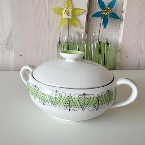 Op de afbeelding: Wit keramisch serveerschaaltje met deksel en twee handvatten. Het schaaltje is versierd met een groen bladerpatroon en een groene rand. Het deksel heeft een kleine, ronde handgreep. Het schaaltje staat op een wit oppervlak.