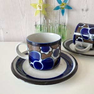 Könnte beinhalten: Zwei Keramik-Kaffeetassen und Untertassen mit blauem Blumenmuster. Die Tassen haben weiße Henkel und stehen auf weißen Untertassen mit blau-braunen Streifen. Der Hintergrund umfasst Glasblumendekorationen.