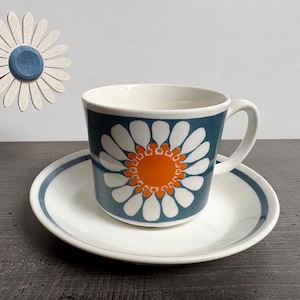Può includere: Un set di tazza e piattino in ceramica con un motivo a margherita. La tazza e il piattino sono bianchi con un bordo blu. La tazza presenta uno sfondo blu navy con un motivo a margherita bianca e un centro arancione. Una margherita decorativa è sullo sfondo.