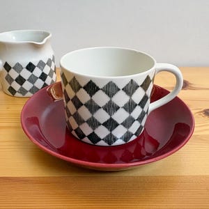 Op de afbeelding: Een witte keramische koffiekop met een zwart-grijs ruitpatroon staat op een rode schotel. Op de achtergrond staat een bijpassende creamer met een handvat en ruitpatroon. De set staat op een houten oppervlak.