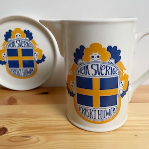 Peut inclure: Un ensemble de pichet et d'assiette en céramique blanche. Les deux pièces présentent un emblème bleu et jaune avec les textes "HEJA SVERIGE" et "FRISKT HUMOR" sur un motif de croix bleu et jaune, représentant le drapeau suédois.