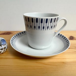 May include: A white teacup and saucer set with a piano key design in navy blue. The cup sits on the saucer, and a small ceramic owl figurine with blue eyes is to the left. The set is on a wooden surface.