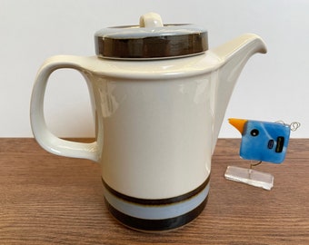 May include: Cream-colored ceramic teapot with a matching lid. The lid and base feature brown and blue stripes. The teapot has a curved handle and a spout. A small, blue glass bird sculpture with an orange beak is next to the teapot.