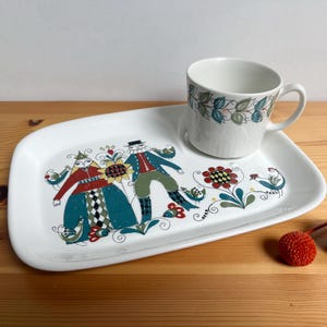 May include: A white ceramic snack set consisting of a rectangular tray and a matching mug. The tray features a colourful illustration of two figures in folk-style clothing, flowers, and birds. The mug has a matching leaf pattern around the rim.