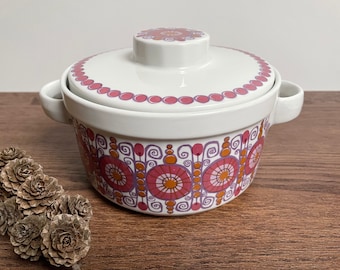 May include: White ceramic casserole dish with a lid, featuring a repeating pattern of pink and orange floral designs. The dish has two small handles and a matching lid with a similar pattern. A decorative piece for serving.