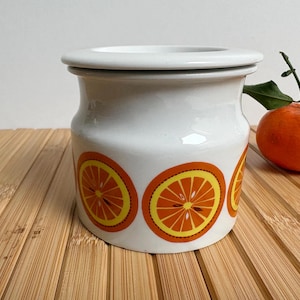 Op de afbeelding: Witte keramische pot met een deksel, versierd met oranje en gele schijfjes citrusvruchten. Een verse sinaasappel met een groene steel ligt naast de pot op een houten oppervlak. De pot heeft een ronde vorm.