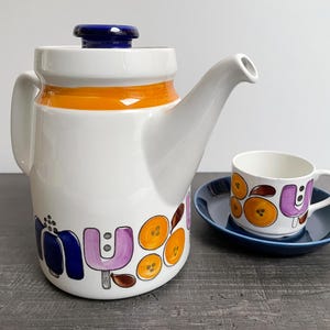 Peut inclure: Une cafetière en céramique blanche avec un couvercle bleu et une bande orange, ainsi qu'une tasse et une soucoupe assorties. La cafetière et la tasse présentent un motif floral coloré bleu, violet et orange. La soucoupe est bleu foncé uni.