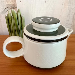 Op de afbeelding: Witte keramische theepot met een donkergroen en zwart deksel en sierlijsten. De theepot heeft een afgeronde body met een textuurpatroon en een grote, gebogen handgreep. Een decoratieve groene plant staat op de achtergrond.