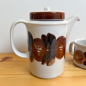 Könnte beinhalten: Eine weiße Keramik-Teekanne mit braunem Deckel und passender Tasse. Die Teekanne und die Tasse sind mit einem sich wiederholenden Blumenmuster in Braun- und Schwarztönen verziert. Die Teekanne hat einen gebogenen Griff und einen Ausguss.