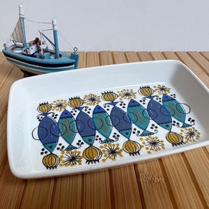 Può includere: Piatto rettangolare in ceramica bianca con un motivo a pesci blu, turchese e gialli. I pesci sono stilizzati con un design moderno della metà del secolo. Una piccola barca a vela giocattolo blu e bianca sullo sfondo.